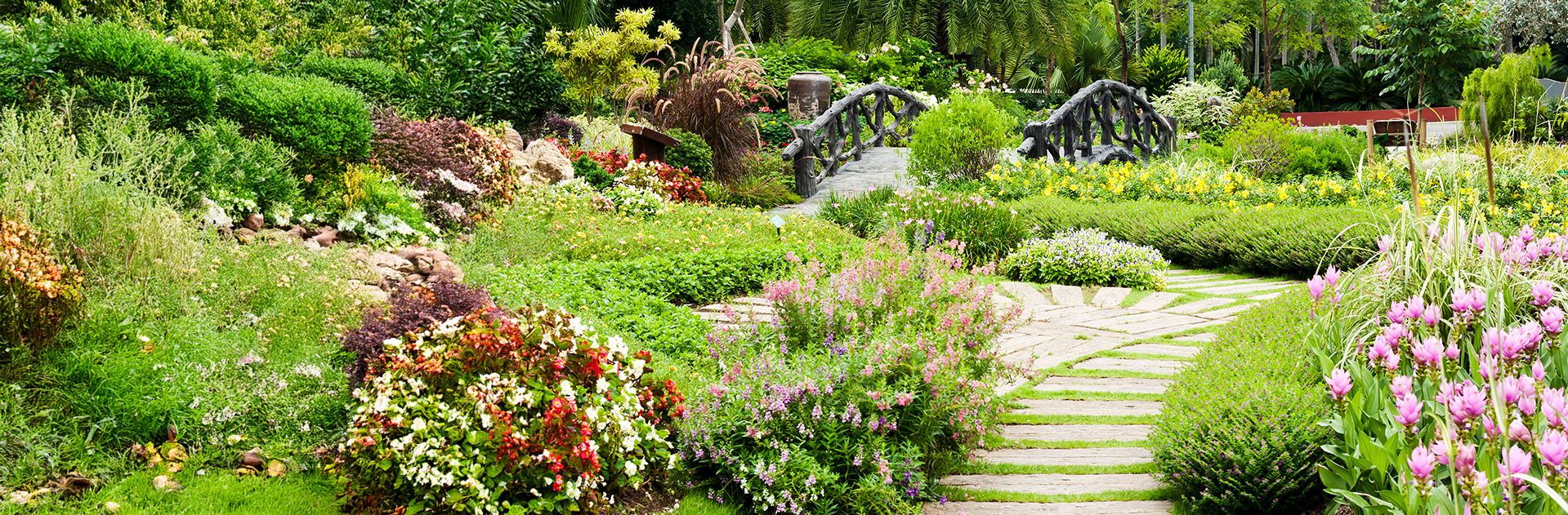 Gepflasterter Weg führt durch einen Garten mit bunten Blumenbeeten und einem kleinen Holzbrückchen im Hintergrund
